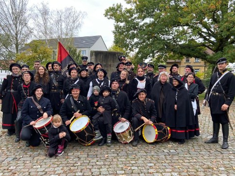 Gruppenbild des Vereins: Erwachsene in schwarzen Uniformen, Frauen in Tracht und Kinder mit Trommeln